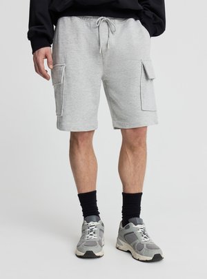 Mężczyzna w jasnoszortach typu cargo ze ściągaczem, czarnych skarpetach i szarych butach sportowych stoi na jednolitym tle.