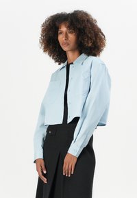Korte lichtblauwe blouse met lange mouwen, knoopsluiting en een borstzak met een hartvormige borduursel. Glad stoftextuur.