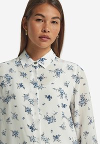 Vit blus med blå blommönster, kragdesign, knappstängning och långa ärmar. Lätt, mjukt tyg med subtil lyster.