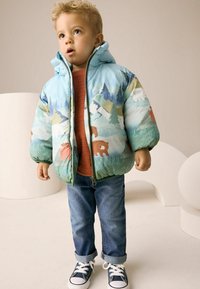 Veste matelassée avec un imprimé animal bleu et vert, mettant en vedette des ours et des montagnes enneigées. Fermeture éclair, associée à une chemise couleur rouille et un jean.