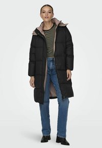 Svart lang puffjakke med lys rosa fôr, med glidelås, sidelommer og hette, båret over en stripete skjorte og jeans.