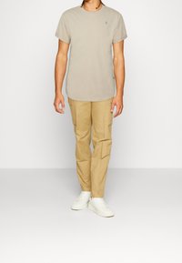 Světle béžové bavlněné tričko s krátkým rukávem, které je kombinováno s hnědými cargo kalhotami a bílými teniskami. Cargo kalhoty mají boční kapsy.