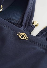 Marineblåt badedragtstof med glat tekstur. Har guldfarvet metal og syninger langs kanterne samt en cirkulær logo.