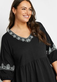 Tunica nera con scollatura a V, caratterizzata da ricami floreali bianchi lungo la scollatura e le maniche, e un corpo ampio e arricciato.