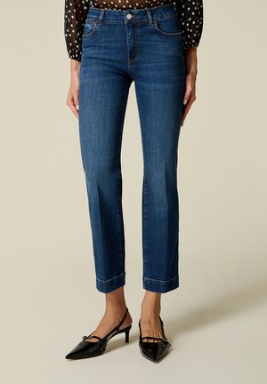 Frau trägt dunkelblaue Jeans mit geradem Bein und schwarze Slingback-Schuhe mit niedrigem Absatz, steht vor einem schlichten Hintergrund.