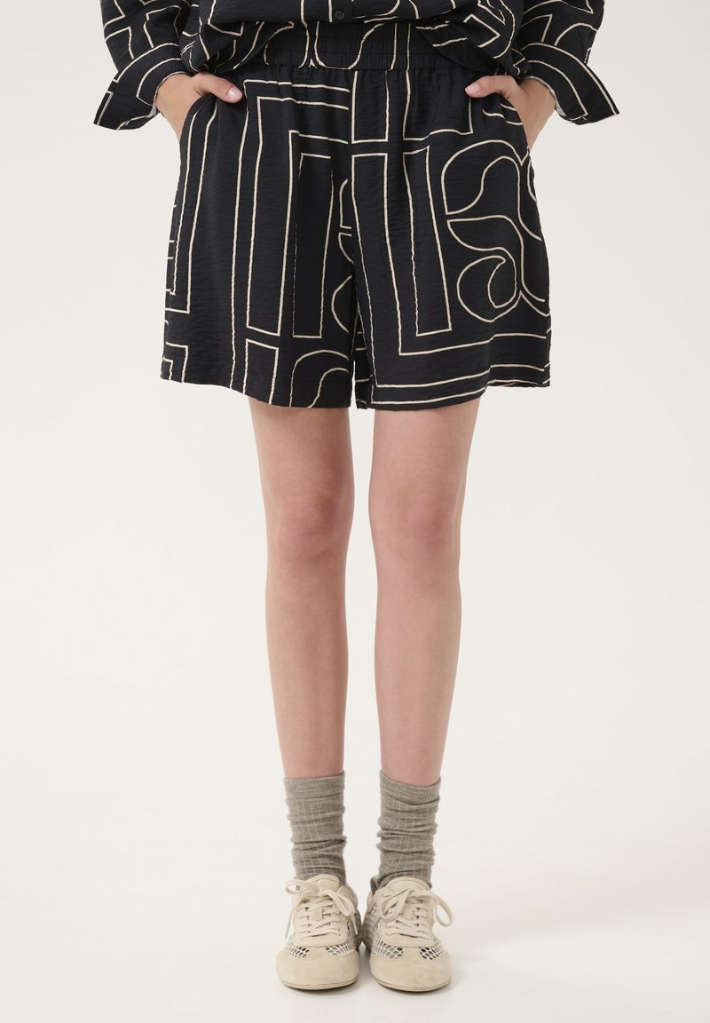 Personne portant un short noir avec des lignes géométriques claires, des chaussettes grises et des baskets beiges, debout les mains dans les poches.