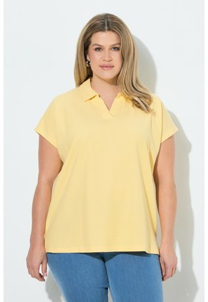 Kobieta o długich blond włosach, ubrana w żółtą koszulę z krótkim rękawem i kołnierzykiem oraz niebieskie dżinsy, stoi na białym tle.