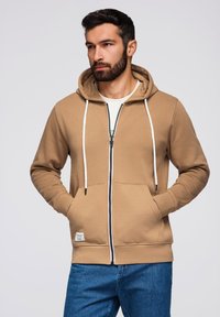 Uomo con barba che indossa un felpa beige con zip e jeans blu, mani nelle tasche, che guarda di lato su uno sfondo grigio uniforme.