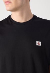 Zwarte katoenen t-shirt met een ronde hals en een klein wit rechthoekig logo-label met rode letters op de linkerkant van de borst.