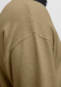 Tan sweater med en tekstureret overflade, der har design med sænkede skuldre og ribbet halsudskæring. Glat strikket materiale, minimale sømme.