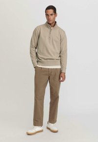 Felpa beige con mezzo zip, polsini a coste, indossata sopra una maglietta bianca. Pantaloni marrone chiaro e sneakers bianche con suola beige.