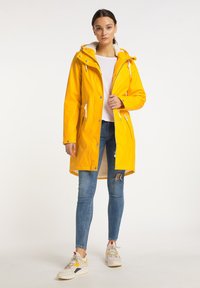 Geel waterdicht jack met een bontgevoerde capuchon, ritssluiting aan de voorkant, zijzakken en verstelbare trekkoorden, gecombineerd met blauwe jeans en sneakers.