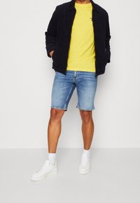 Chaqueta Navy, camiseta amarilla, pantalones cortos de denim azul. Zapatillas blancas con suela texturizada y calcetines de canalé. Conjunto informal con un ajuste relajado.