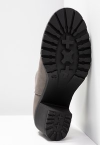 Semelle d'une botte grise avec une profonde semelle noire présentant des motifs en croix et en X, inclinée contre un mur d'angle blanc.