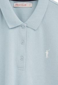 Hellblaues Polohemd mit drei Knöpfen, klassischem Kragen, kleinem weißem, gesticktem Polo-Spieler-Logo auf der Brust und "POLO CLUB"-Label innen am Kragen.