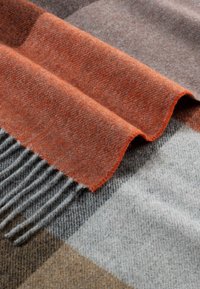 Weicher, gewebter Wollschal mit orange, grau und braunem Karomuster, gefaltet, um die strukturierte Fransenkante und das Stoffdetail zu zeigen.