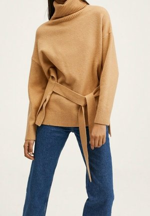 Kvinde iført en kamel-farvet rullekravesweater med bånd i taljen, kombineret med blå denimjeans mod en ensfarvet baggrund.