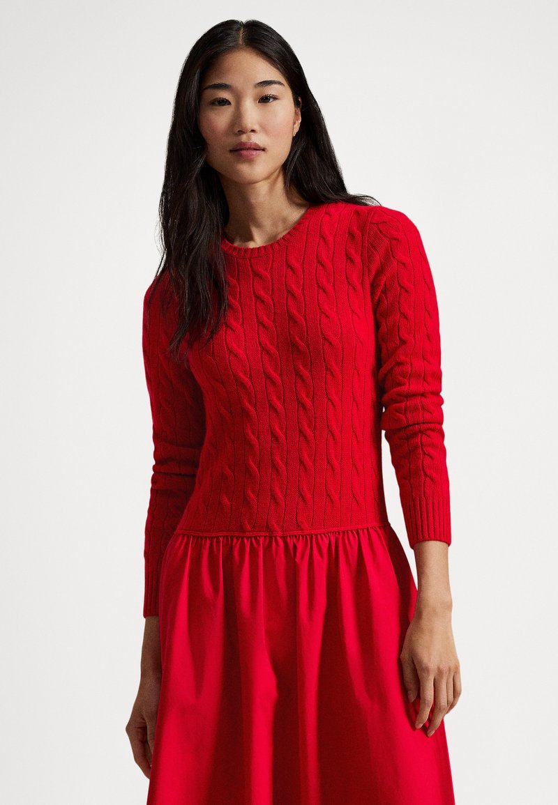 Rdeč pulover z vezenjem v kitah, okrogel izrez in dolgi rokavi, v kombinaciji z nabrano rdečo krilco, ki ustvarja učinek večplastne obleke.