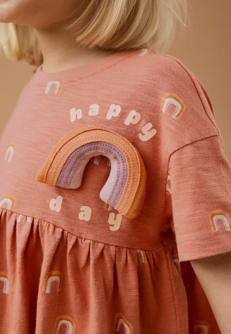 Jeune enfant portant une robe pêche avec des motifs arc-en-ciel et un patch arc-en-ciel en tissu, texte « journée heureuse » sur la poitrine.