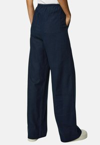 Wijde broek in een marineblauwe stof met een elastische tailleband, voorzien van twee achterzakken en een gladde textuur. In combinatie met witte schoenen.