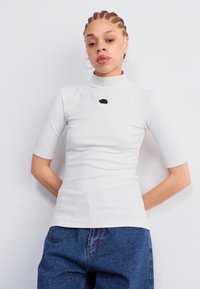 Balta trumpa rankovėmis marškinėlių su mock turtleneck apykakle tekstūruota medžiaga ir maža juoda logotipas krūtinėje, derinama su laisvomis mėlynomis džinsinėmis kelnėmis.