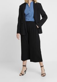 Zwarte maatpakblazer over een blauwe overhemD, gecombineerd met zwarte wijde broek en zwarte sandaaltjes met hoge hakken. Glad textiel overal.
