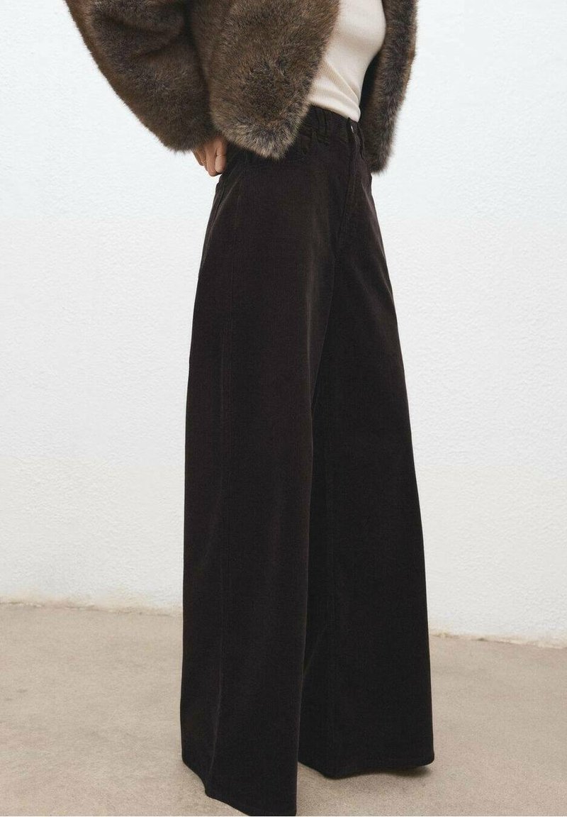 Personne portant un pantalon noir à jambes larges, un haut blanc et une veste marron en fourrure, debout contre un mur blanc uni et un sol beige.