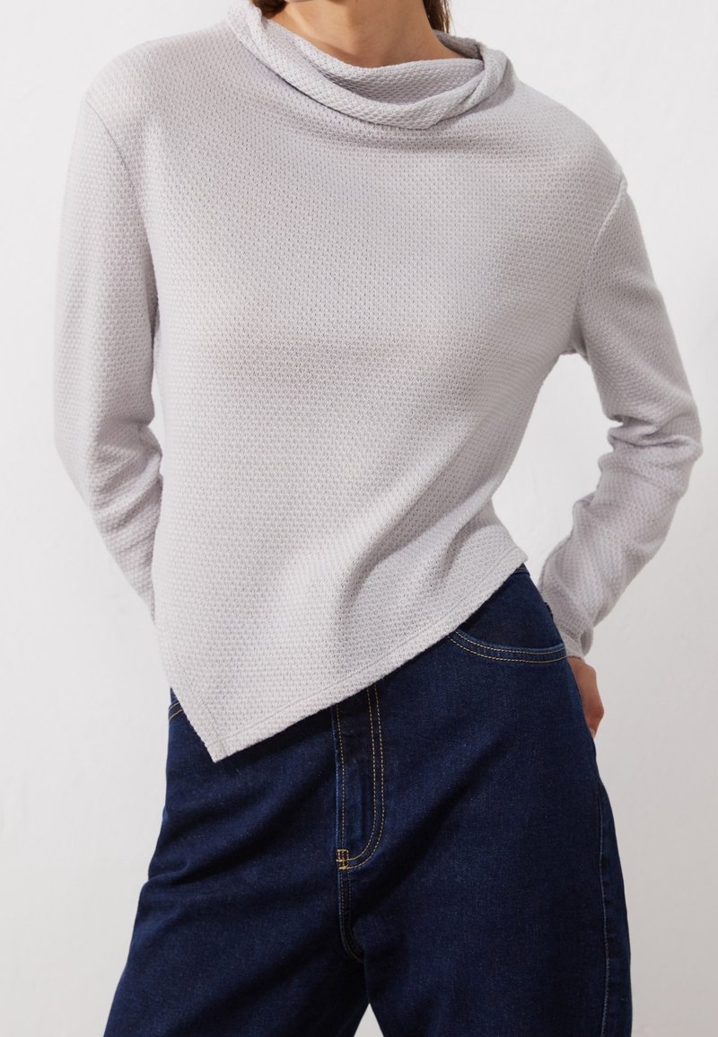 Femme portant un haut gris clair texturé à manches longues avec un ourlet asymétrique et un jean bleu foncé, les mains derrière le dos.