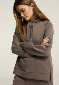 Brązowa dziewiarska bluza z kapturem o luźnym kroju, z teksturowanej tkaniny, z regulowanym sznurkiem i bocznymi rozcięciami dla wygody i stylu.