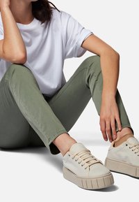 Zapatillas de lona beige con suela de goma texturizada, puntera redonda y cordones planos. Llevadas con pantalones verde olivo y una camiseta blanca.