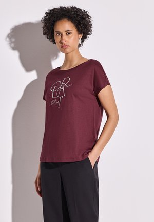 T-shirt en coton bordeaux avec des manches courtes, ornée d'un motif gris clair. Associé à un pantalon noir. Texture lisse, coupe décontractée.