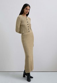 Compleu din tricot bej format dintr-o jachetă scurtă cu nasturi negri și o fustă lungă cu un model texturat în zigzag. Botine negre cu toc.