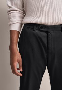 Osoba w jasnobeżowym, prążkowanym swetrze z podwiniętymi rękawami oraz czarnych spodniach z guzikami, z ręką luźno opartą o bok na jednolitym tle.