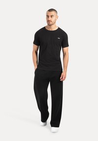 Man met kort haar, die een zwart T-shirt, losse zwarte broek en witte sneakers draagt, staande met zijn handen in de zakken op een witte achtergrond.