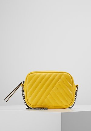 Sac bandoulière rectangulaire matelassé jaune avec bandoulière chaîne noire et tirette de fermeture éclair sur plateforme blanche sur fond gris.