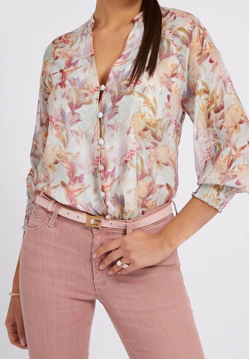 Blumenbluse aus transparenter Stoff mit Knopfdetails, in sanften Rosa- und Grüntönen. Kombiniert mit hellrosa, enganliegenden Jeans und einem schmalen Gürtel.