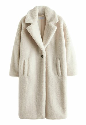 Personne portant un long manteau teddy crème, un pull en maille marron, un pantalon marron brillant et des baskets blanches marchant sur un trottoir en ville.