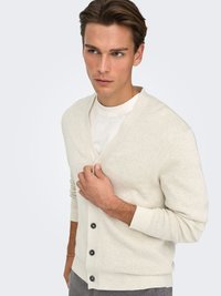 Cardigan beige clair en texture tricotée, avec un col en V, des manches longues et six boutons foncés sur le devant.