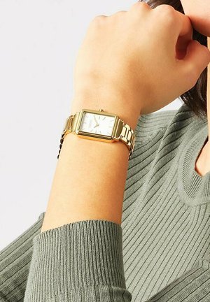 Goldenes rechteckiges Uhr mit einem Metallarmband, weißem Zifferblatt, silbernen Stundenmarkierungen und einer kleinen Krone an der Seite. Trägt an einer Hand gegen einen grünen Pullover.