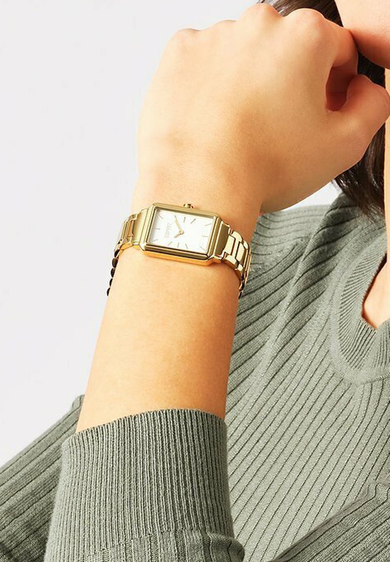 Orologio rettangolare dorato con bracciale a maglie metalliche, quadrante bianco, indicatori dell'ora argentati e una piccola corona sul lato. Indossato su una mano contro un maglione verde.