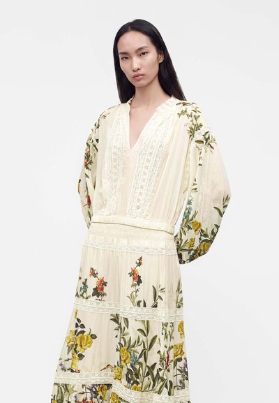 Femme aux longs cheveux noirs portant une robe crème à imprimé floral, avec des détails en dentelle et des manches longues bouffantes sur un fond uni.