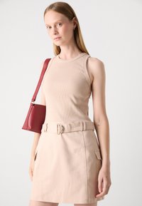 Débardeur beige à côtes associé à une mini-jupe beige clair à ceinture avec des poches latérales. Le mannequin porte un petit sac à main rouge.