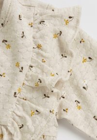 Haut beige à motifs floraux avec de petites fleurs jaunes et des accents marron. Tissu texturé avec des volants le long de l'encolure et des manches.