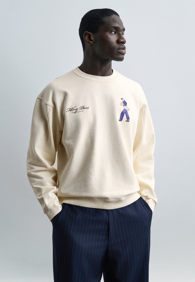 Άνδρας με τα χέρια στις τσέπες, φορ άει εκρού φούτερ με μικρή φιγούρα που περπατά και κείμενο, συνδυασμένο με μπλε σκούρο παντελόνι ριγέ, σε απλό φόντο.