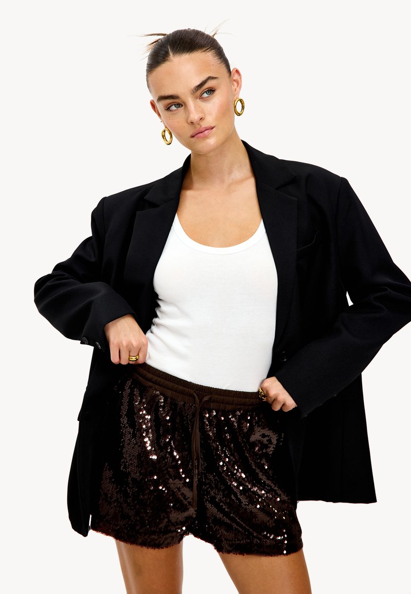 Blazer noir oversize, débardeur blanc et short en sequins marron avec un waistband élastique. Boucles d'oreilles en or visibles. Design simple et moderne.