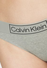 Sivi bombažni bikini z raztegljivim materialom in črnim blagovno znamko pasom, ki prikazuje ime "Calvin Klein" v pravokotnem oblikovanju etikete.