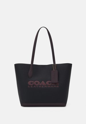 Czarna skórzana torba typu tote z ciemnobrązowymi uchwytami, wytłoczonym logo "COACH LEATHERWARE" oraz wiszącą skórzaną przypinką przymocowaną złotym łańcuszkiem.