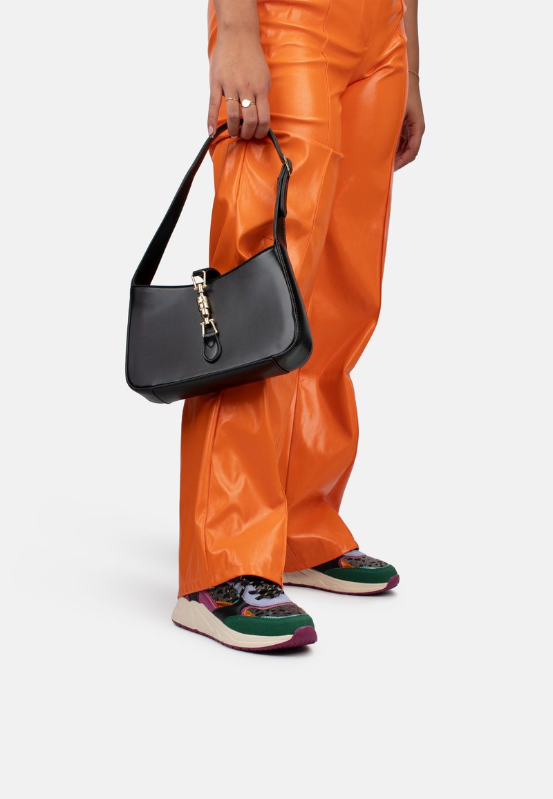 Bolso de cuero negro de forma rectangular, con un acento de hardware dorado. Textura suave, sostenido por una mano contra pantalones naranjas.