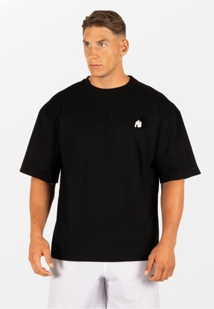 Man draagt een zwart oversized T-shirt met een klein wit gorilla-logo op de borst, gecombineerd met een witte korte broek, kijkt iets naar rechts.