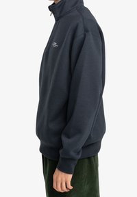 Granatowy sweter z wysokim kołnierzem, wykonany z miękkiego materiału, z prążkowanymi mankietami i detalami logotypu na przodzie. Widok z boku podkreśla luźny krój.